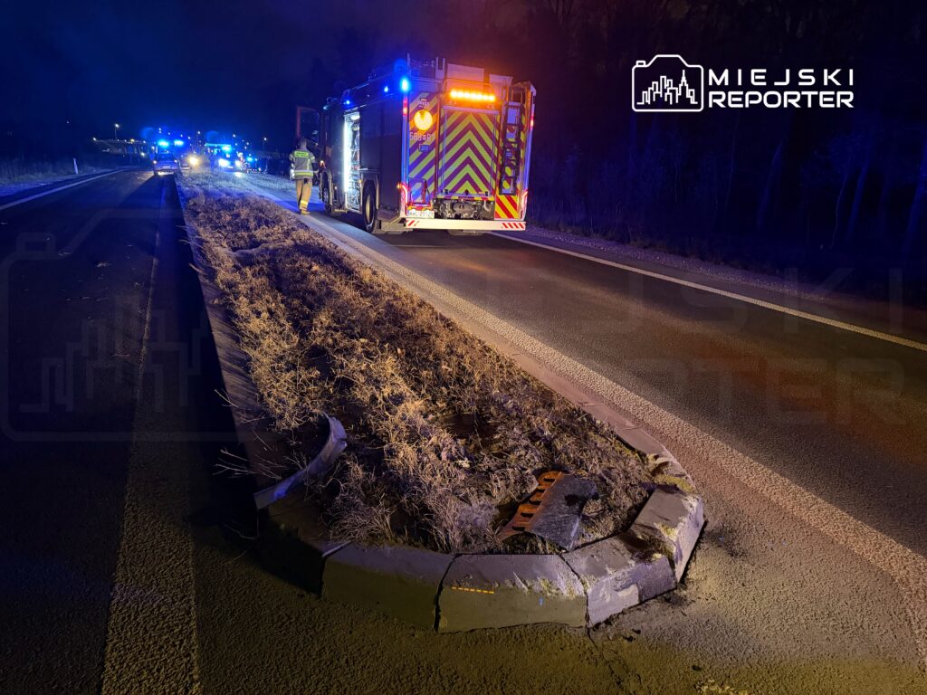 Na drodze w nocy, strażacy przy wozie strażackim z włączonymi sygnałami, otoczeni przez policyjne pojazdy.