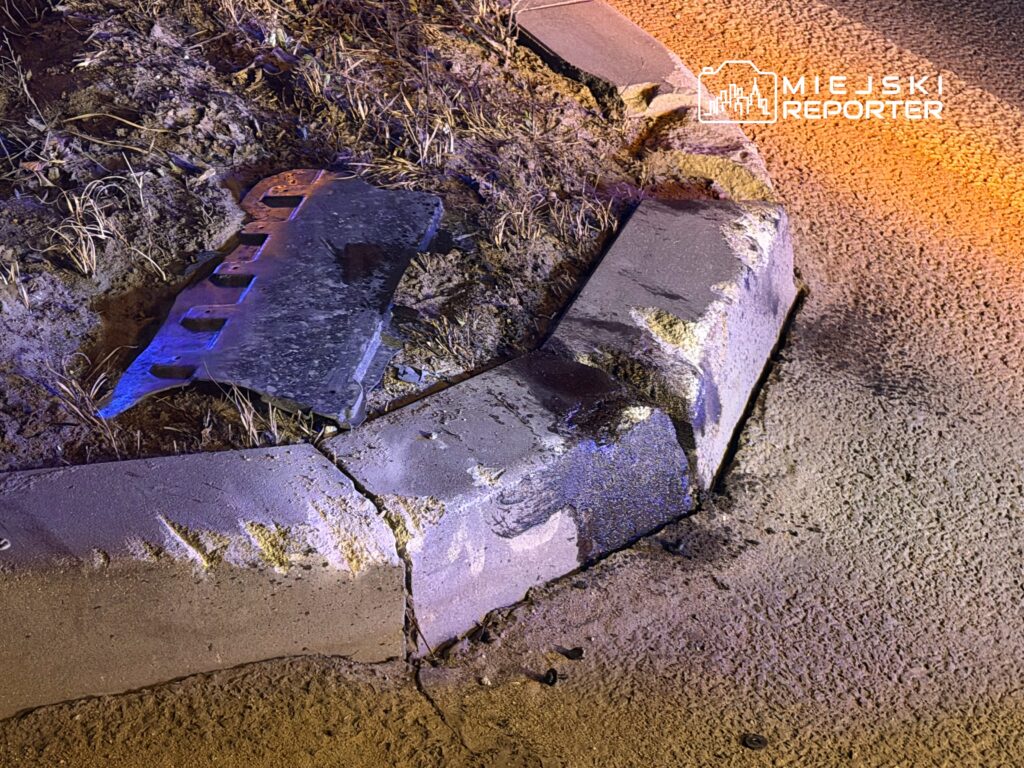 Uszkodzona krawężnik z widocznymi śladami uszkodzeń oraz odłamkiem czarnego elementu leżącym obok.