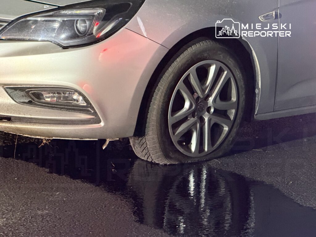 Samochód osobowy z uszkodzoną oponą zaparkowany na mokrej nawierzchni, obok widoczne refleksy światła.