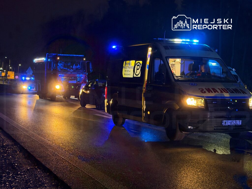 Na drodze nocą zatrzymane pojazdy służb ratunkowych z włączonymi sygnałami świetlnymi.