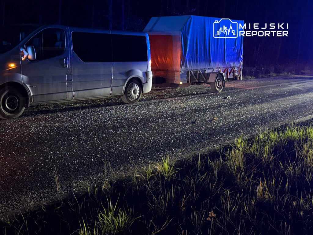 Srebrny van z przyczepą stoi na drodze w nocy, otoczony przez trawę, z widocznymi reflektorami.