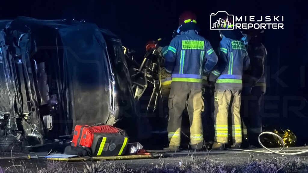 Grupa strażaków w mundurach z odblaskami prowadzi działania ratunkowe przy przewróconym samochodzie w nocy.