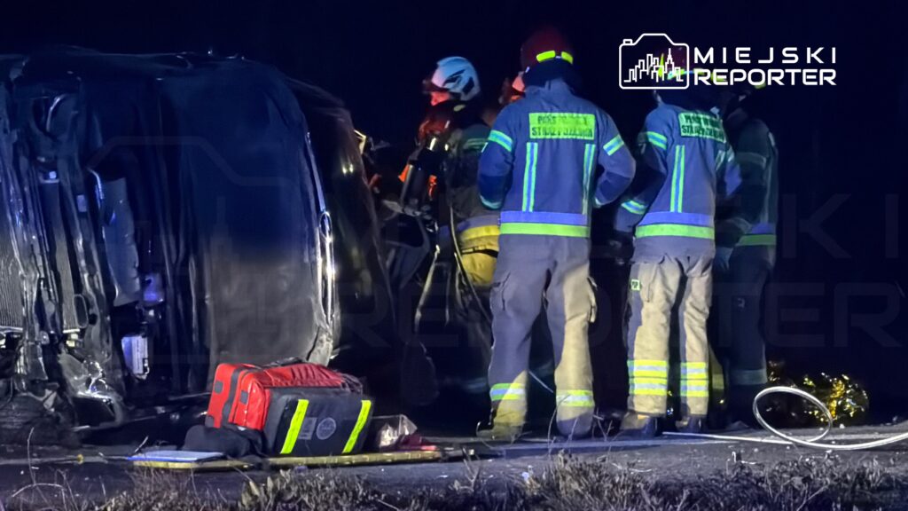 Grupa strażaków w mundurach z odblaskami pracuje przy przewróconym pojeździe na drodze nocą.