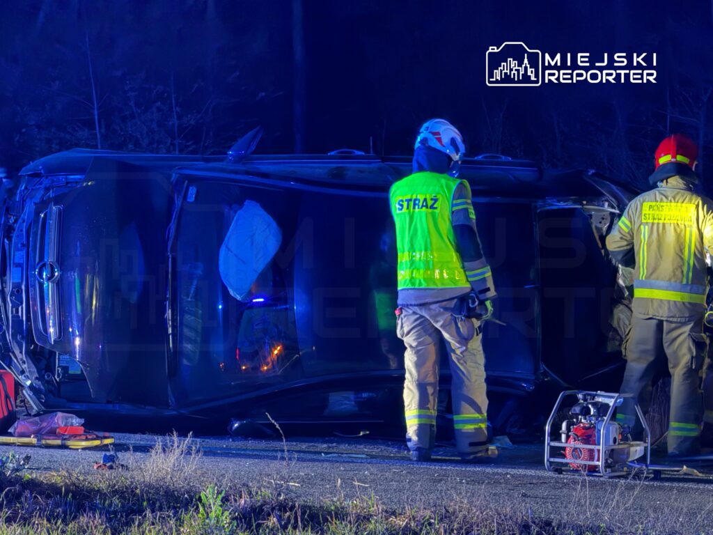 Ratownicy w odblaskowych kamizelkach pracują przy przewróconym samochodzie na poboczu drogi, w nocy.