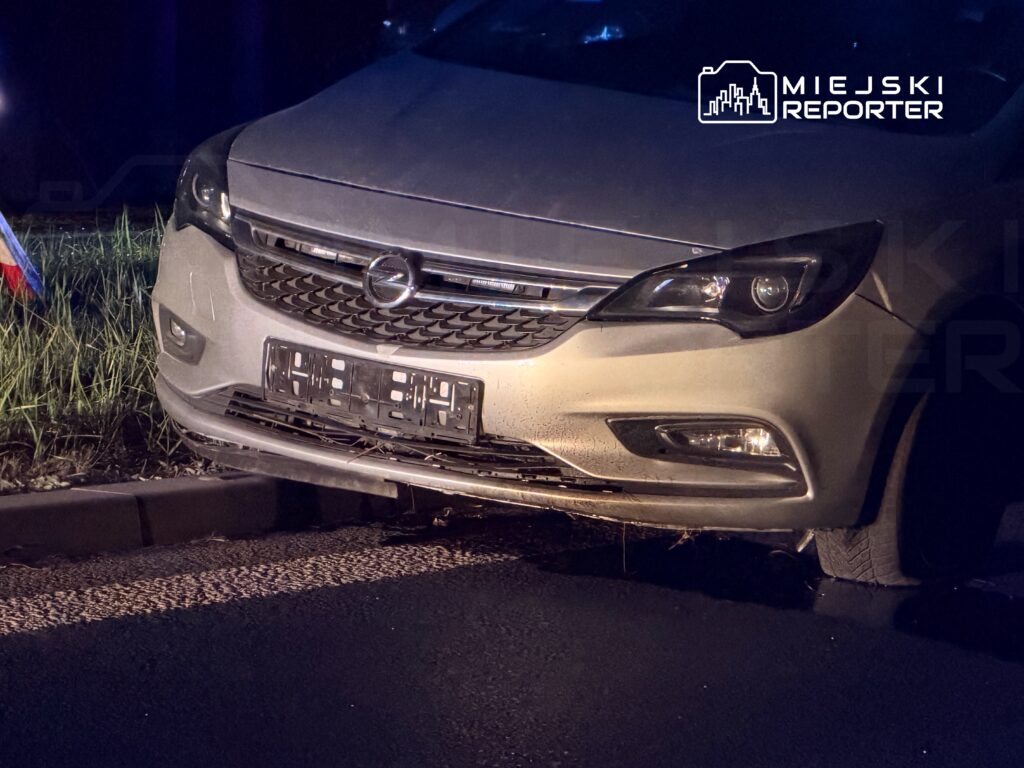 Uszkodzony samochód osobowy zaparkowany na poboczu drogi, z widocznymi zarysowaniami na masce i zderzaku.