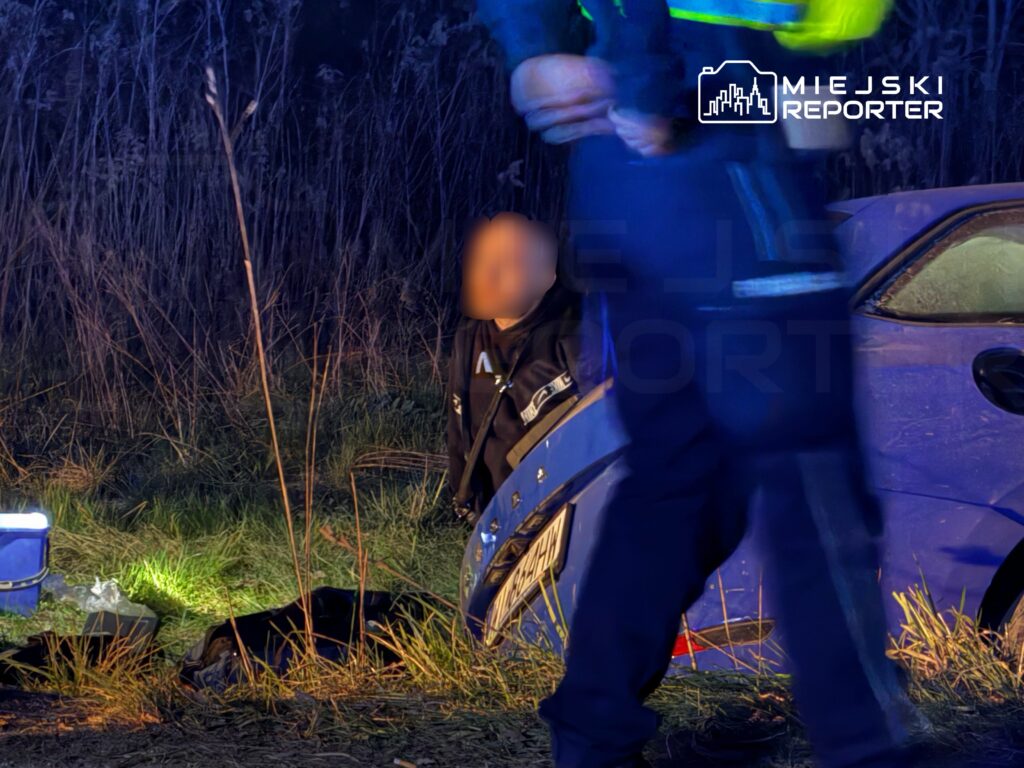 Mężczyzna w ciemnym ubraniu siedzi na trawie, podczas gdy funkcjonariusz policji go przeszukuje w ciemnym otoczeniu.