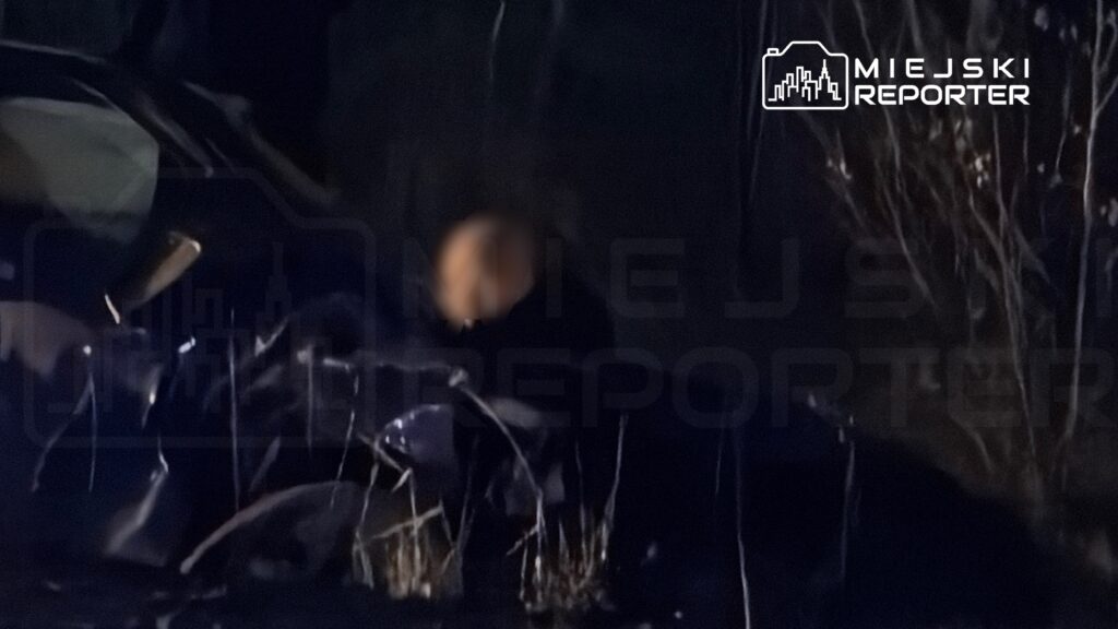 Mężczyzna w ciemnym ubraniu przeszukuje teren w nocy, otoczony przez zarośla i ciemne tło.