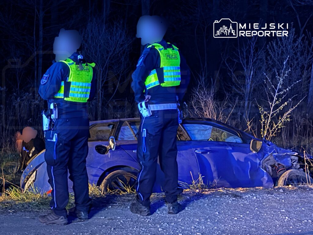 Dwaj policjanci w odblaskowych kamizelkach stoją obok uszkodzonego samochodu na poboczu drogi, w ciemnym, leśnym otoczeniu.
