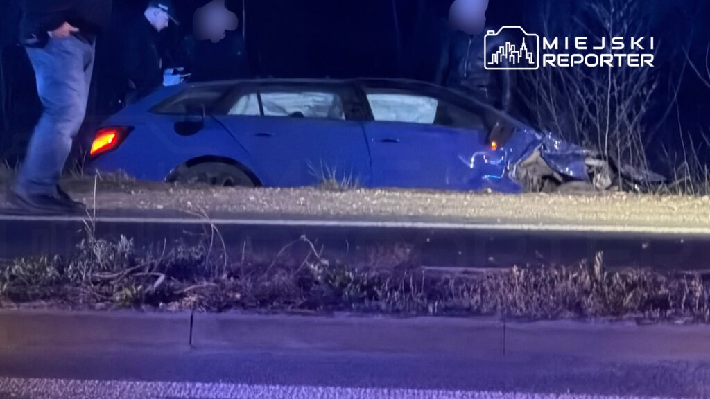 Nieoznakowany niebieski samochód osobowy z uszkodzeniami, zaparkowany na poboczu drogi, obok mężczyźni w ciemnych ubraniach.