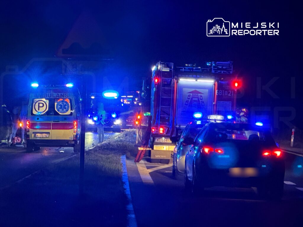 Na drodze nocą stoją pojazdy służb ratunkowych, w tym karetka i wóz strażacki, z włączonymi niebieskimi sygnalizacjami.