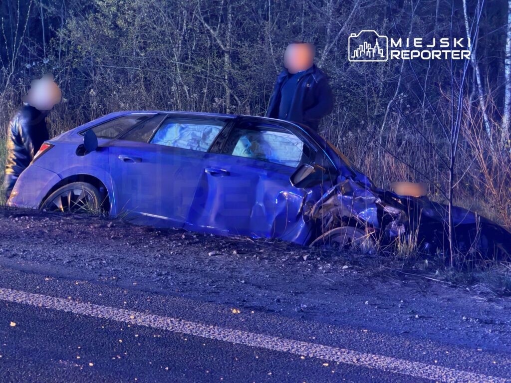 Osobnicy w ciemnych ubraniach oglądają uszkodzony niebieski samochód, który wpadł do rowu przy drodze.