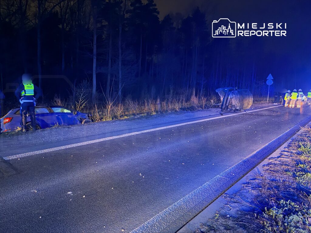Policjant zabezpiecza miejsce wypadku, obok przewrócone auto na poboczu drogi w lesie, w tle błyskające światła.