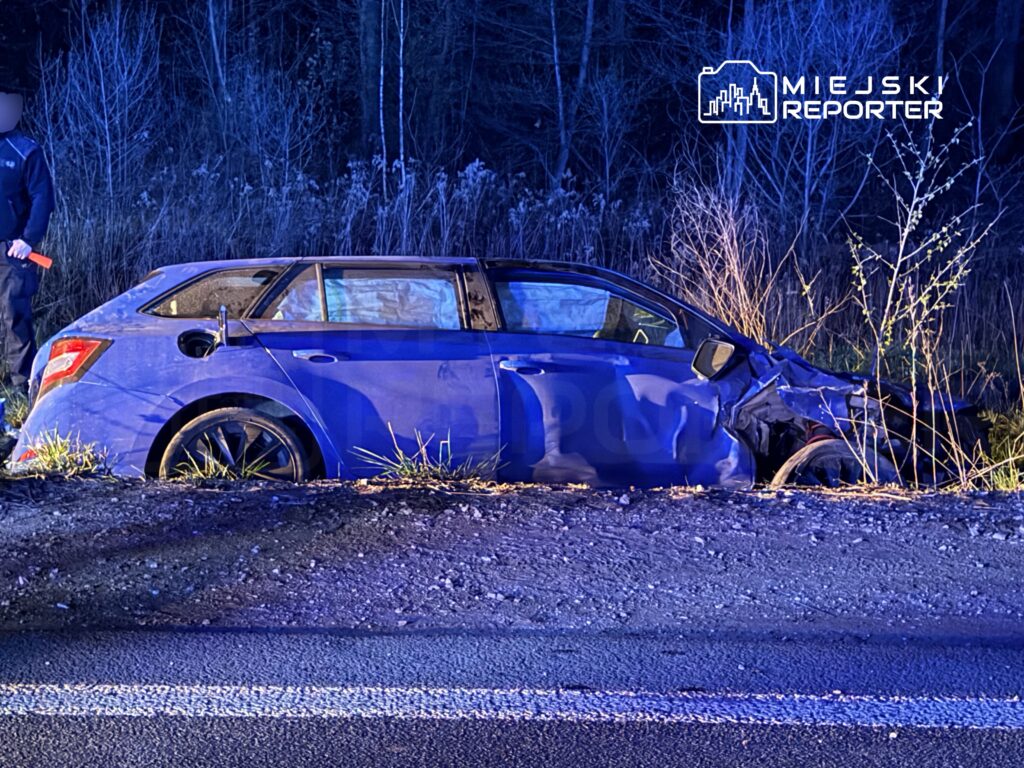 Mężczyzna w mundurze policyjnym bada uszkodzony niebieski samochód osobowy na poboczu drogi.