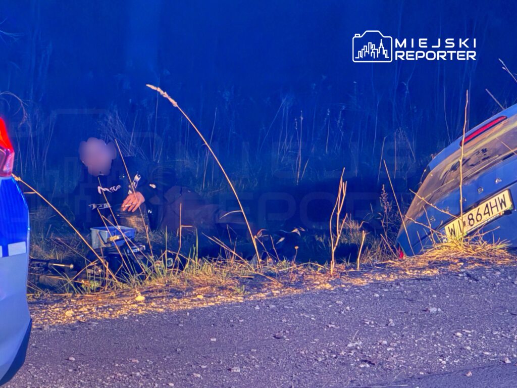 Mężczyzna w mundurze leży obok przewróconego samochodu na poboczu drogi, w otoczeniu wysokiej trawy.