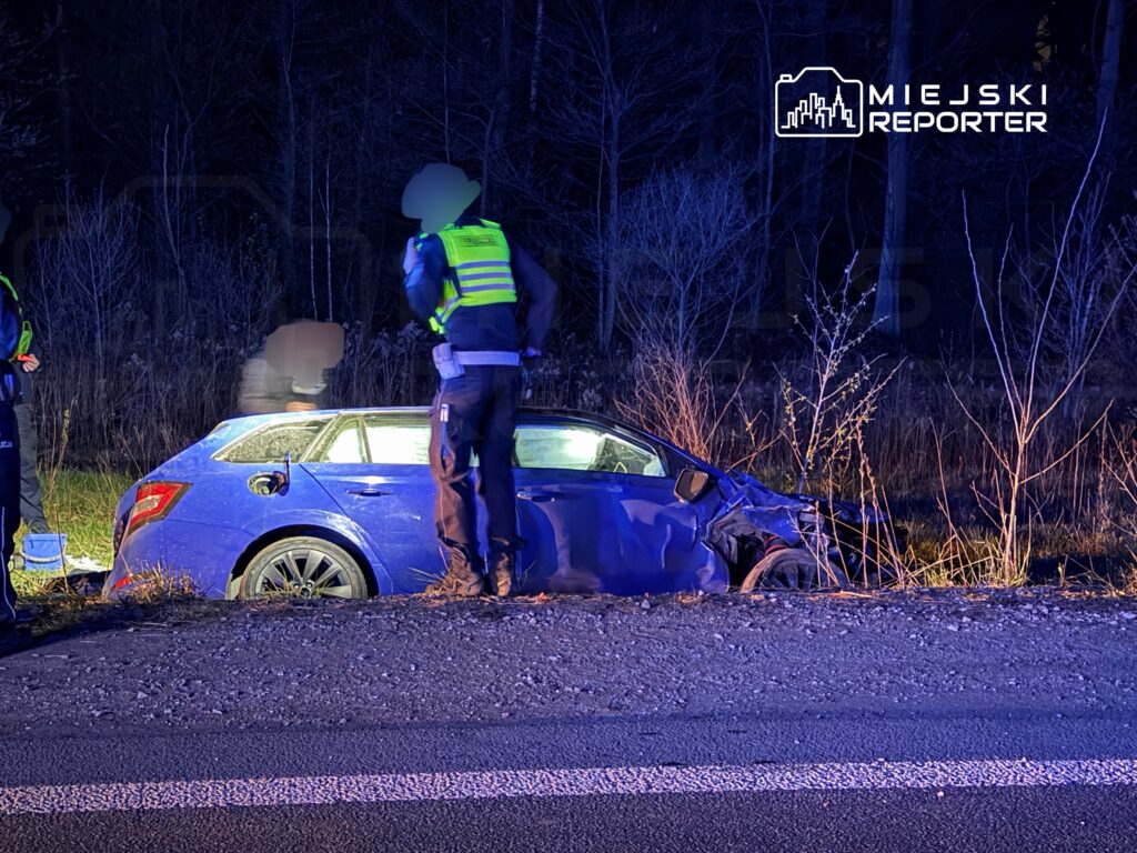 Dwaj policjanci prowadzą czynności na poboczu drogi obok uszkodzonego niebieskiego samochodu osobowego.
