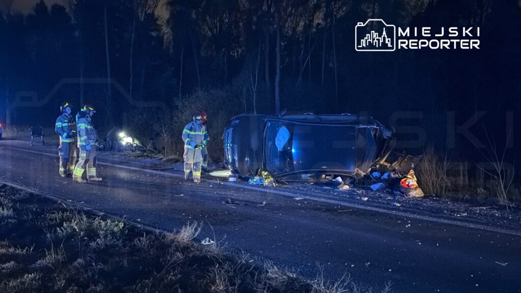 Grupa strażaków w mundurach z latarkami bada przewrócony samochód na drodze w lesie w nocy.