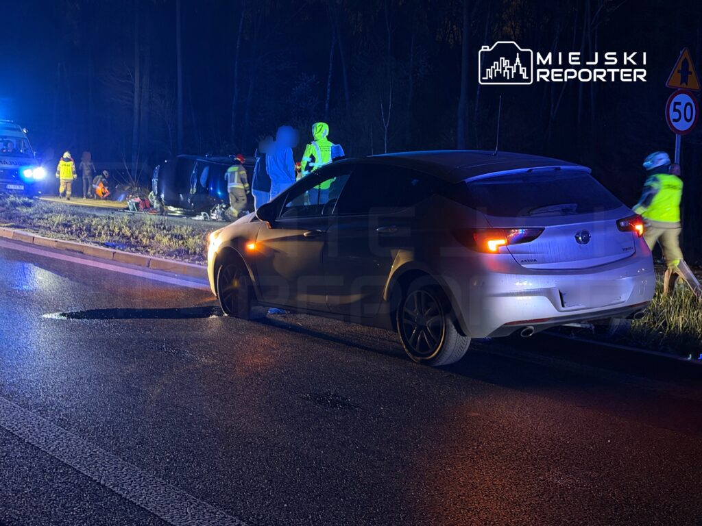 W nocy na drodze leży przewrócony samochód, a obok pracują służby ratunkowe w odblaskowych kamizelkach.