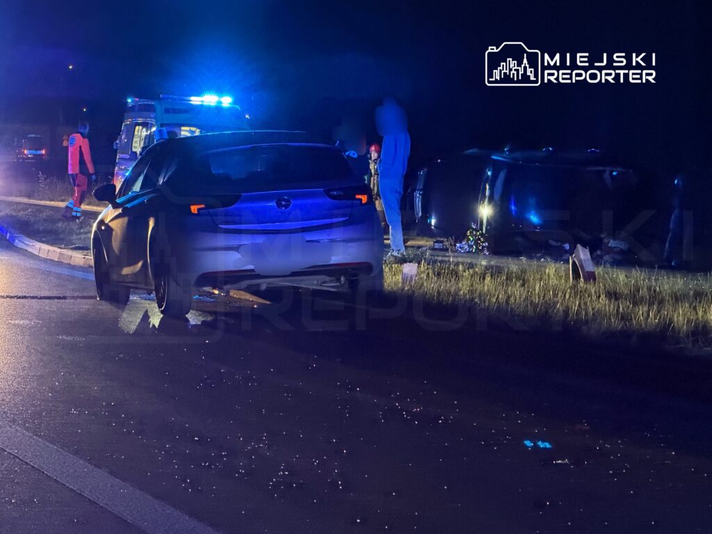 Czarny samochód osobowy stoi na poboczu drogi, obok przewrócony pojazd, a w tle widać karetki pogotowia z włączonymi sygnałami świetlnymi.