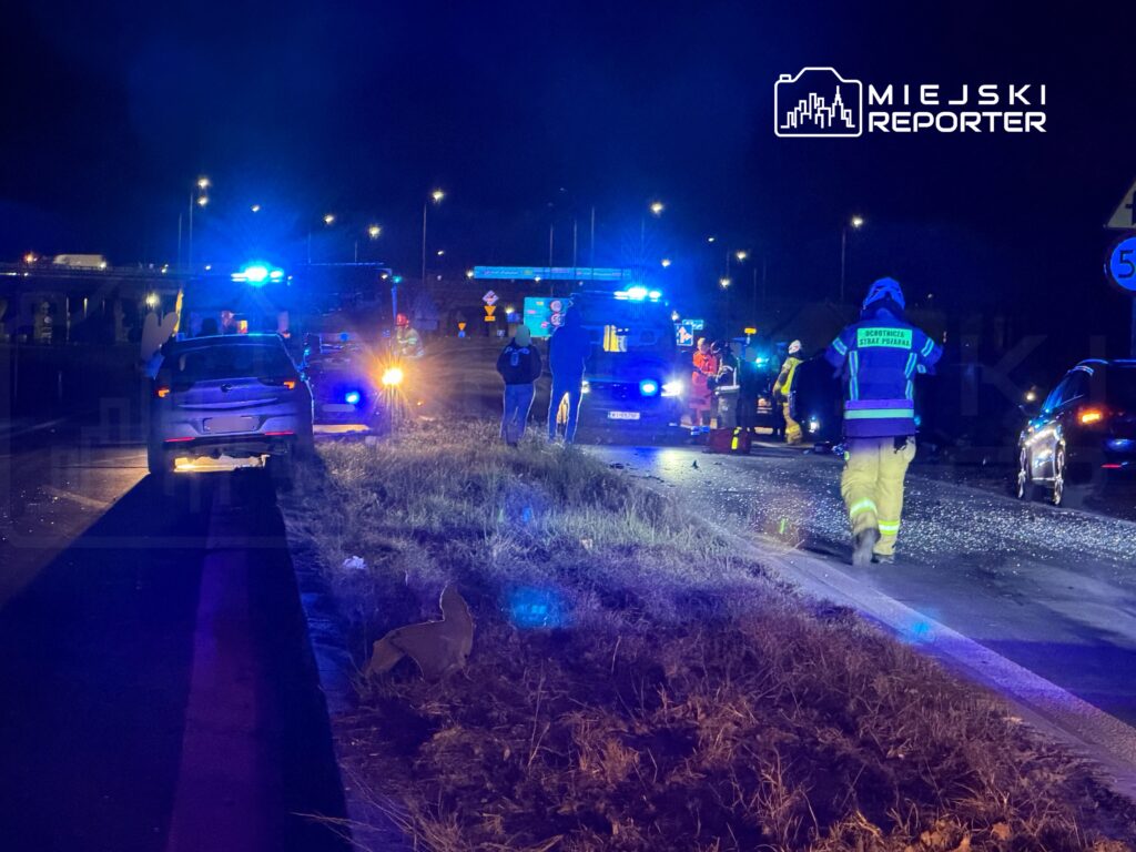 Na drodze w nocy, służby ratunkowe pracują przy wypadku, otoczone niebieskimi światłami sygnalizacyjnymi.