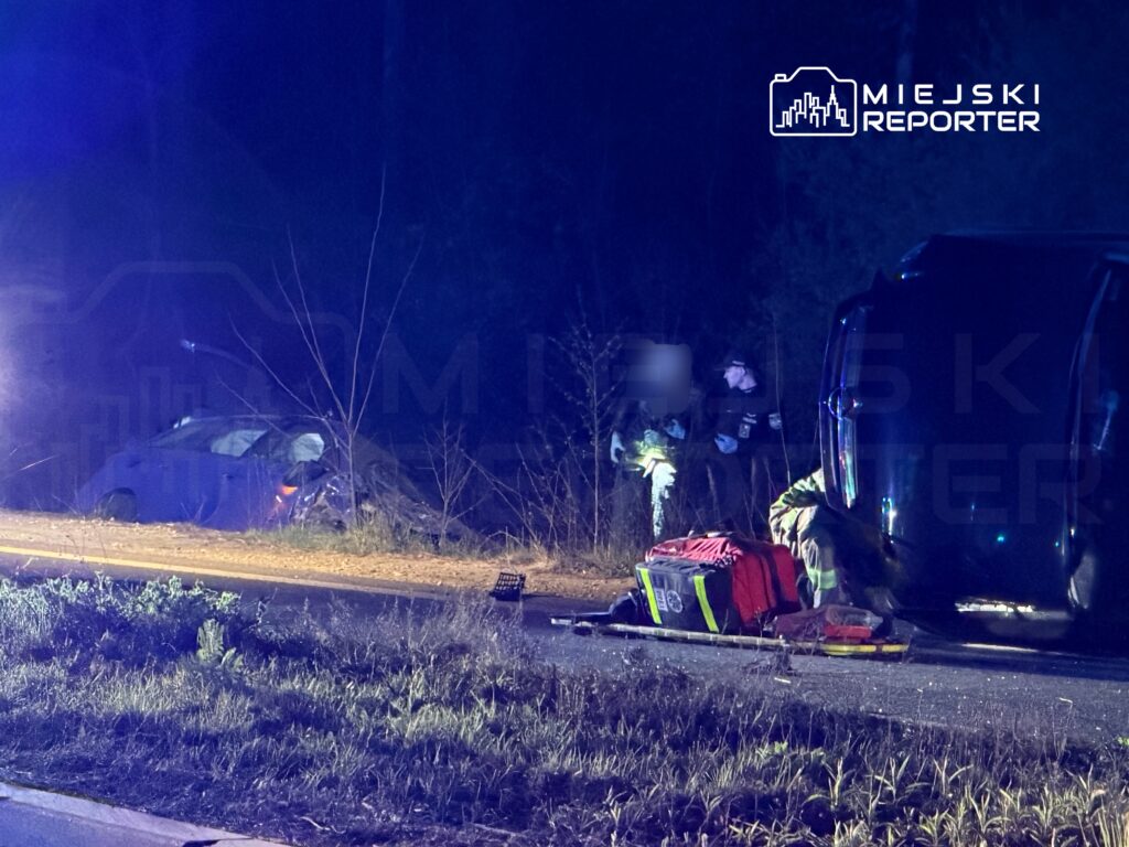 Na drodze w lesie, strażacy udzielają pomocy poszkodowanemu przy przewróconym samochodzie.