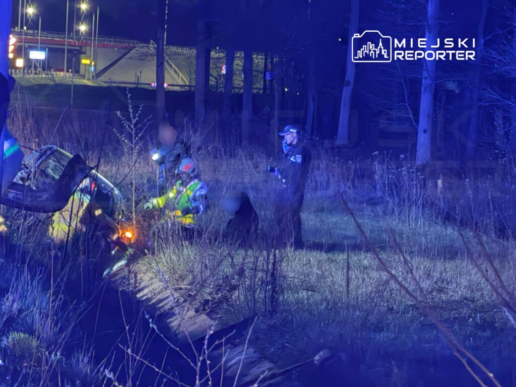 Grupa ratowników w żółtych kurtkach bada przewrócony samochód w lesie, w tle widoczne światła uliczne.