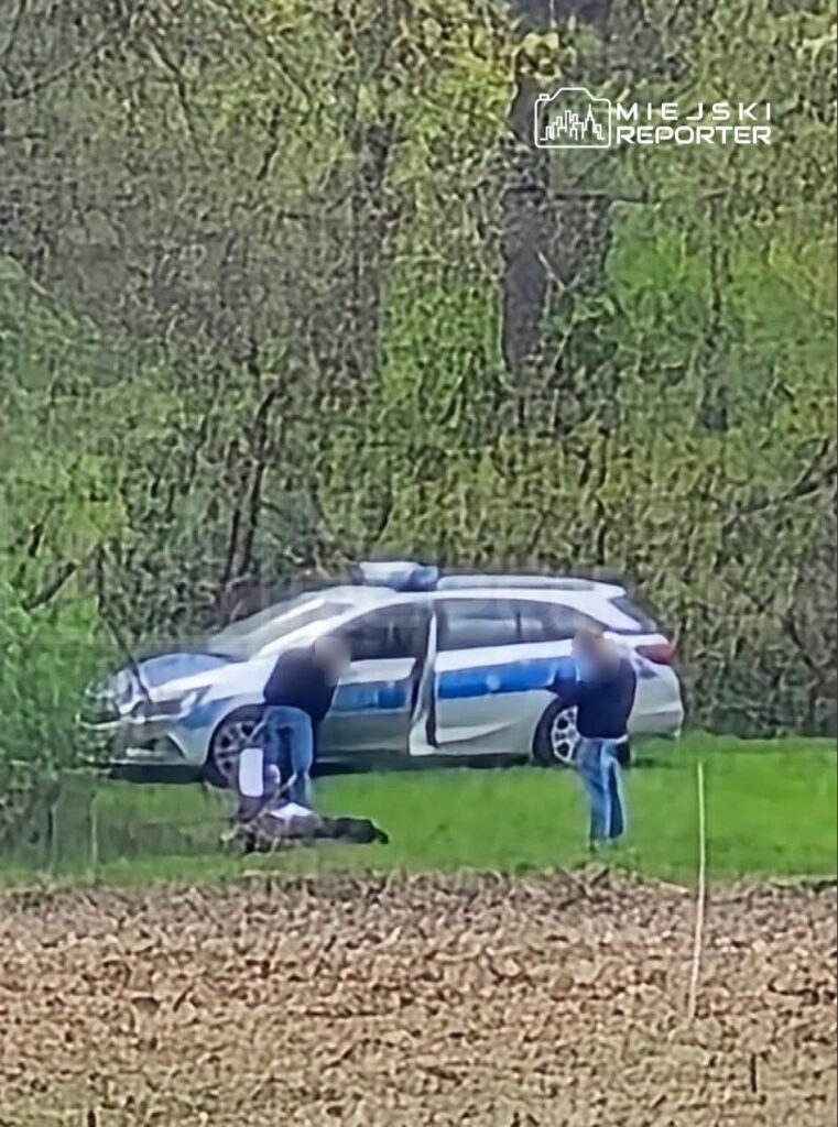 Trzech mężczyzn w dżinsach i ciemnych bluzach pochyla się nad leżącą osobą obok radiowozu w terenie.