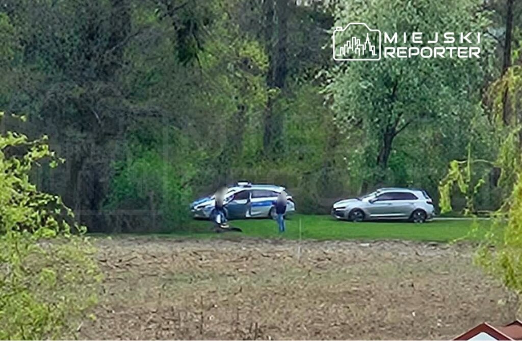 Dwóch mężczyzn rozmawia obok policyjnego samochodu na tle zadrzewionego terenu.