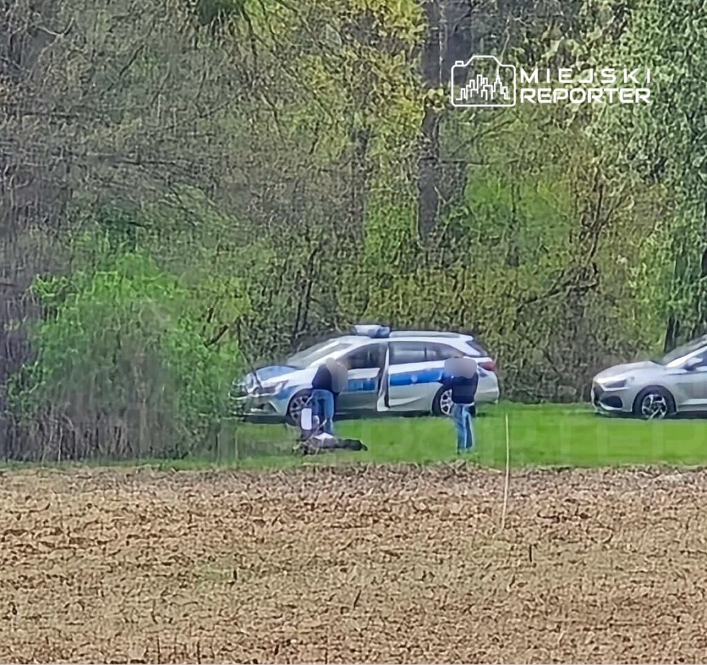 Grupa osób bada miejsce zdarzenia obok policyjnego samochodu na tle zieleni i otwartego pola.