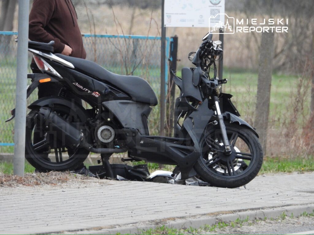 Mężczyzna w brązowej bluzie stoi obok czarnego skuteru zaparkowanego na chodniku w otoczeniu zieleni.