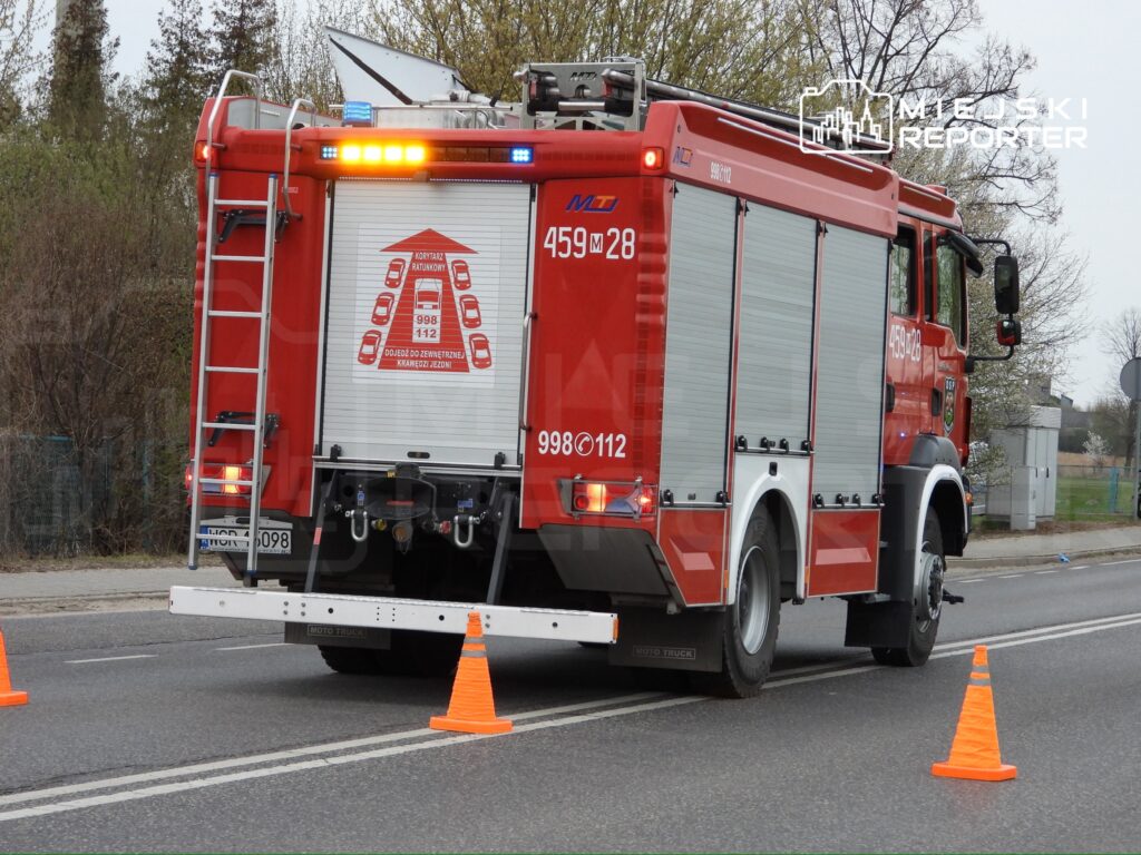 Czerwony wóz strażacki z włączonymi sygnalizatorami zatrzymany na drodze, otoczony pomarańczowymi pachołkami.