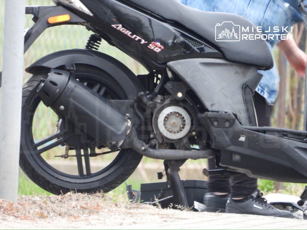 Czarna skuter stoi na chodniku, obok widoczna ręka osoby w niebieskiej kurtce, która dokonuje jego naprawy.