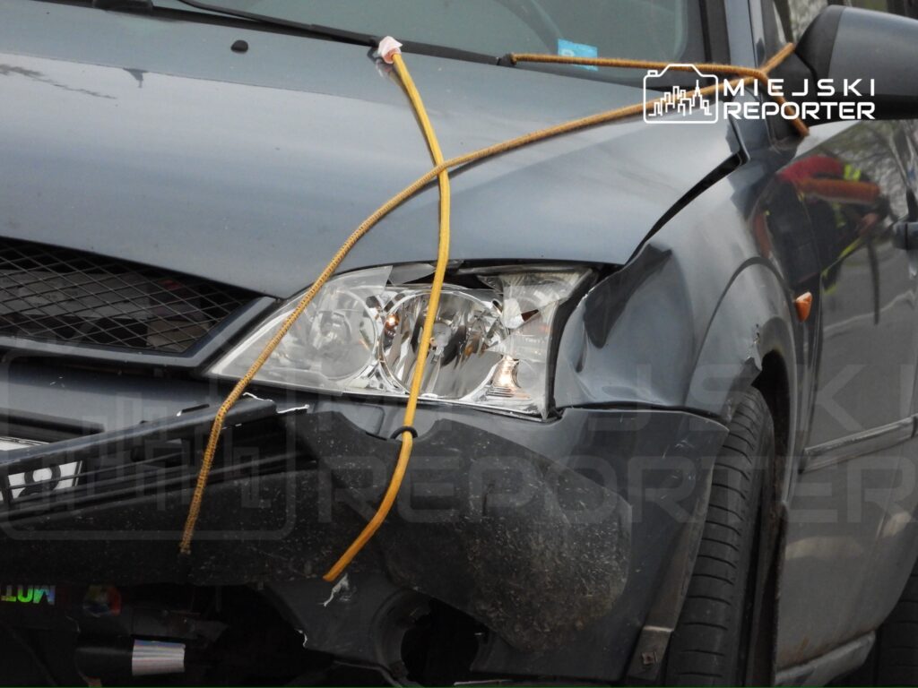 Uszkodzony samochód osobowy z zarysowanym przodem, związaną liną na masce, zaparkowany na ulicy.