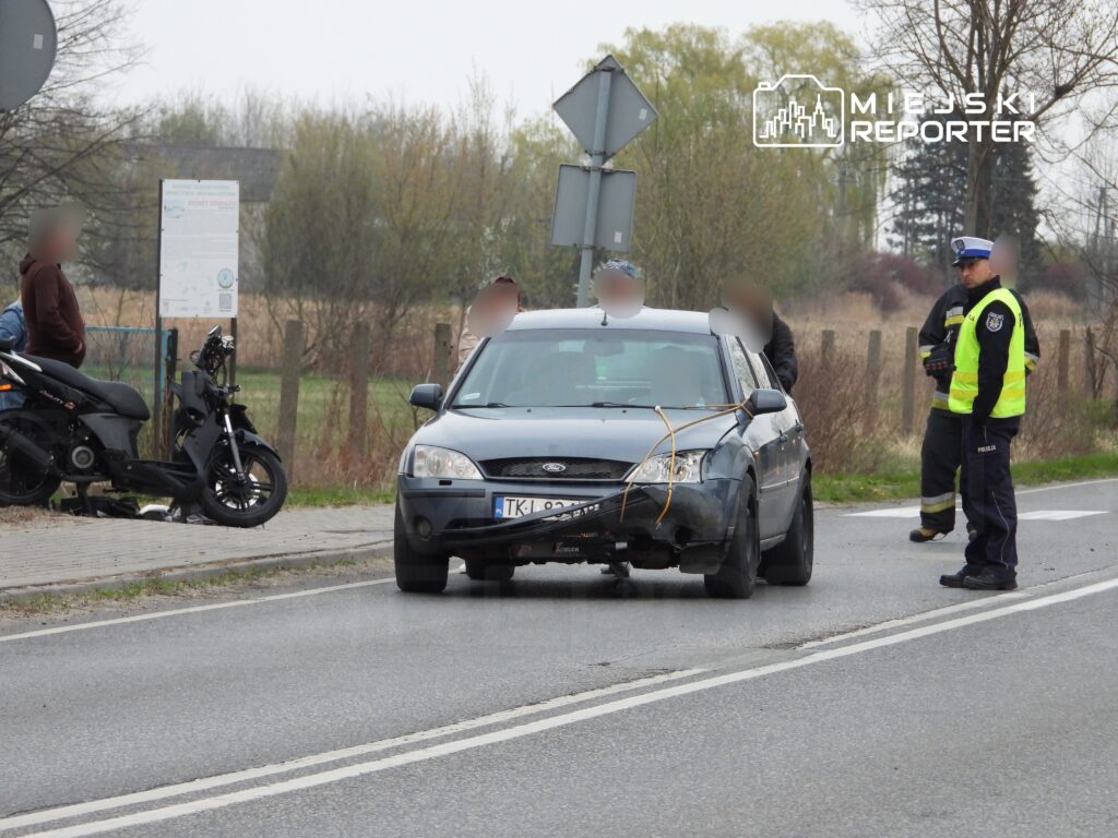 Czarny samochód osobowy z uszkodzeniami stoi na drodze, obok grupka osób oraz policjant w kamizelce odblaskowej.