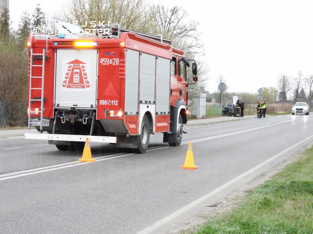 Wóz strażacki z włączonymi sygnałami stoi przy drodze, a strażacy udzielają pomocy poszkodowanemu motocykliście.