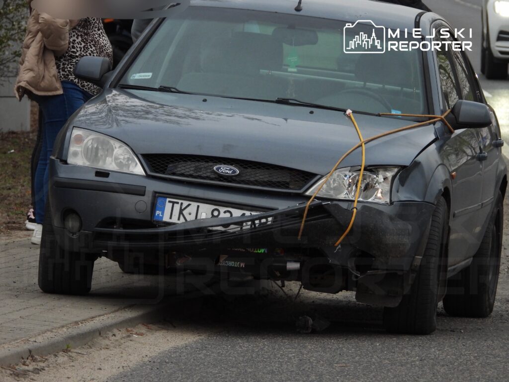 Uszkodzony czarny samochód osobowy z unieruchomionym przodem stoi na poboczu, obok rozmawiające kobiety.