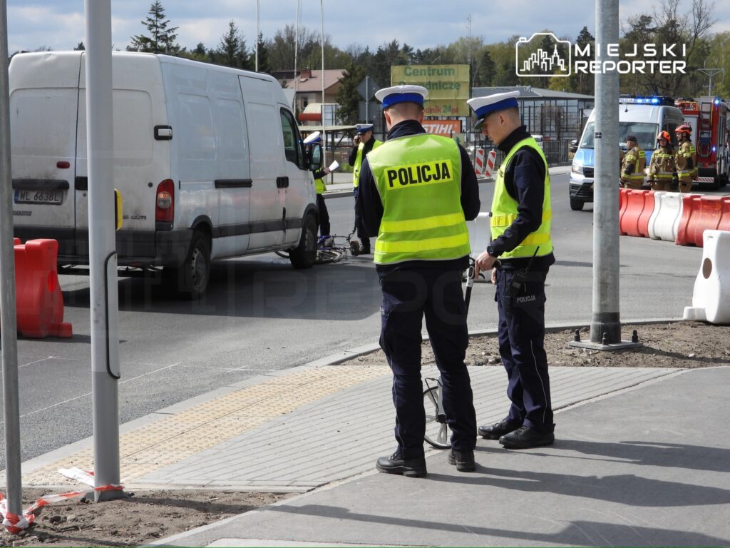 Dwaj policjanci w kamizelkach odblaskowych prowadzą czynności na drodze obok białego dostawczego auta.