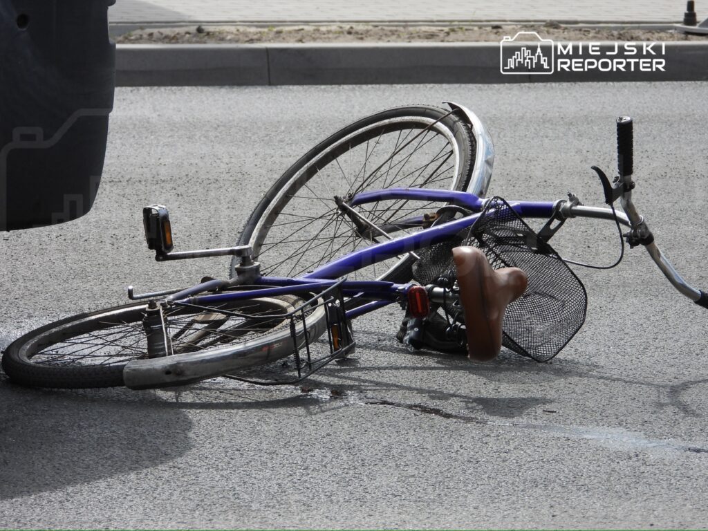 Złamany niebieski rower leży na jezdni, z uszkodzonym przednim kołem i odwróconym koszykiem.