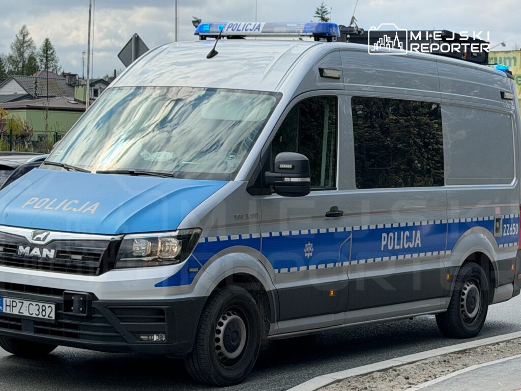 Policyjny pojazd marki MAN zaparkowany na drodze, z włączonymi sygnalizatorami świetlnymi.