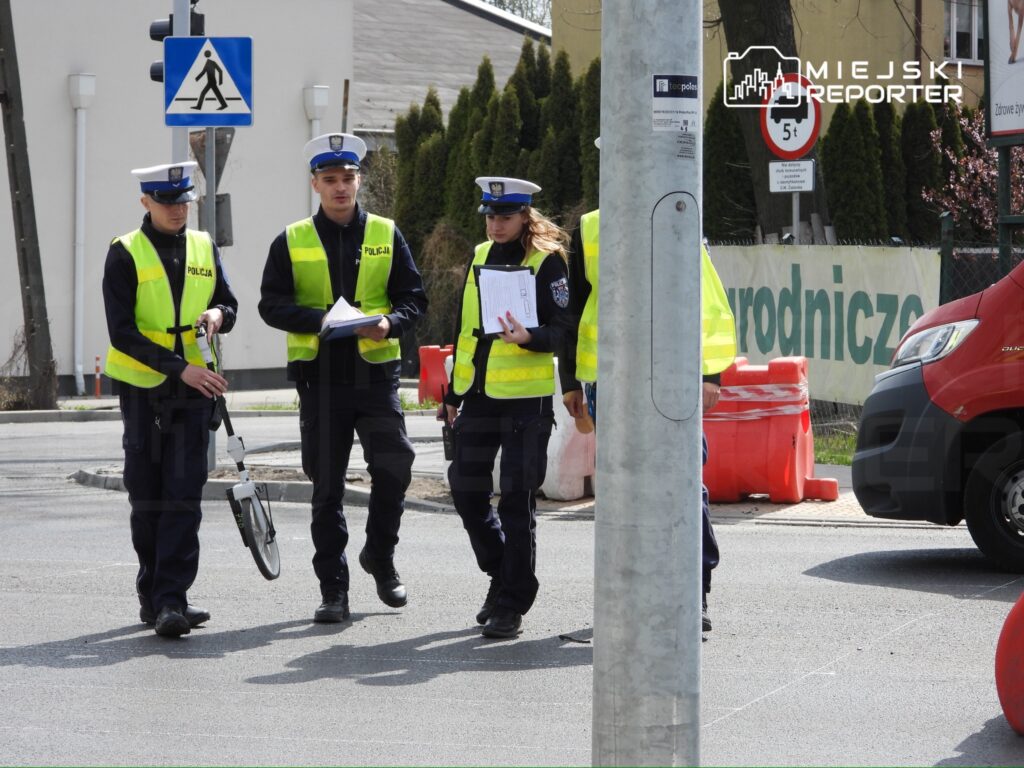 Czterech policjantów w kamizelkach odblaskowych rozmawia na ulicy, trzymając dokumenty i sprzęt pomiarowy.