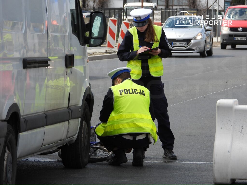 Policjant w kamizelce odblaskowej bada uszkodzenie roweru obok białego furgonu, a towarzysząca mu policjantka spisuje notatki.