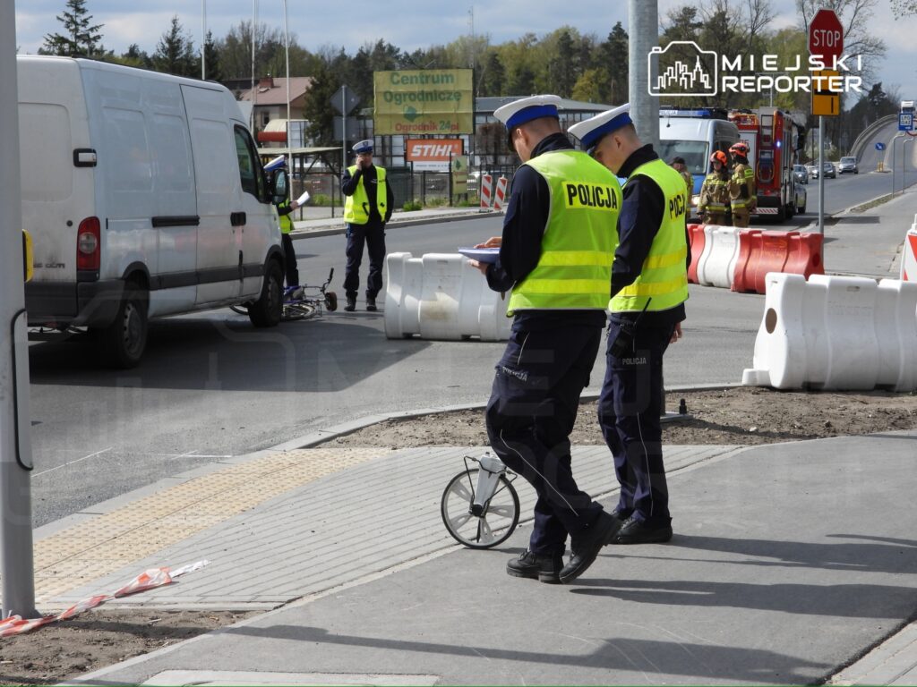 Dwaj policjanci w kamizelkach odblaskowych prowadzą czynności na drodze obok przewróconego roweru.