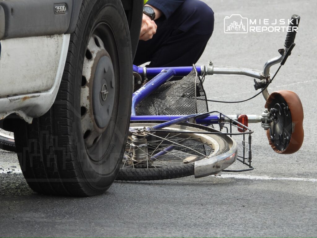Rower leży na asfalcie obok kółka samochodu, a obok stoi mężczyzna sprawdzający uszkodzenia.