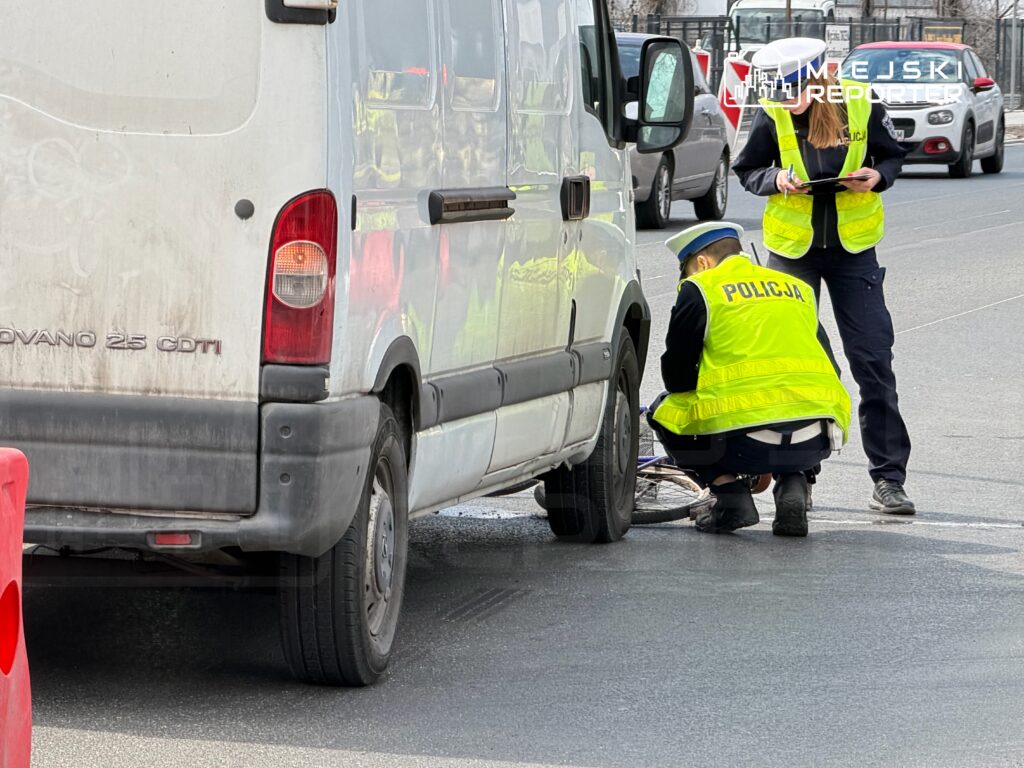 Dwóch policjantów w kamizelkach odblaskowych bada uszkodzone koło w białym samochodzie dostawczym na ulicy.