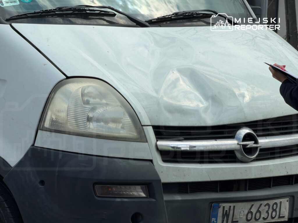 Uszkodzony przód białego vana z wgnieceniem na masce, obok stoi osoba notująca na notatniku.