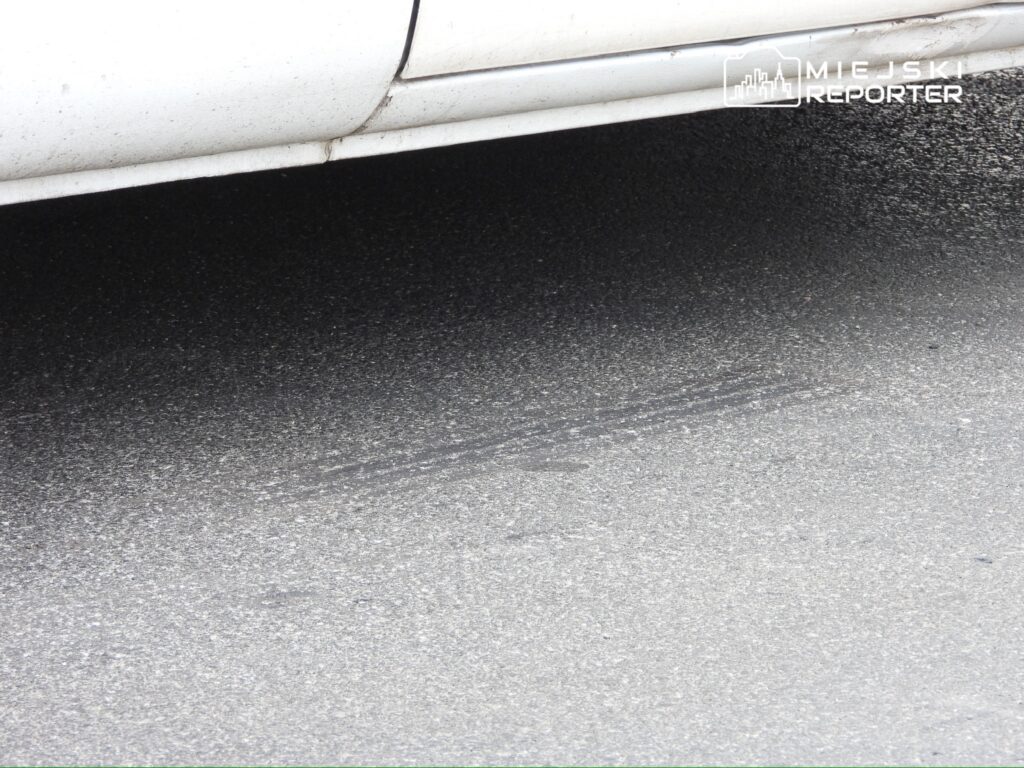 Biały samochód osobowy zaparkowany na szarej nawierzchni, z widocznymi plamami brudu na karoserii.