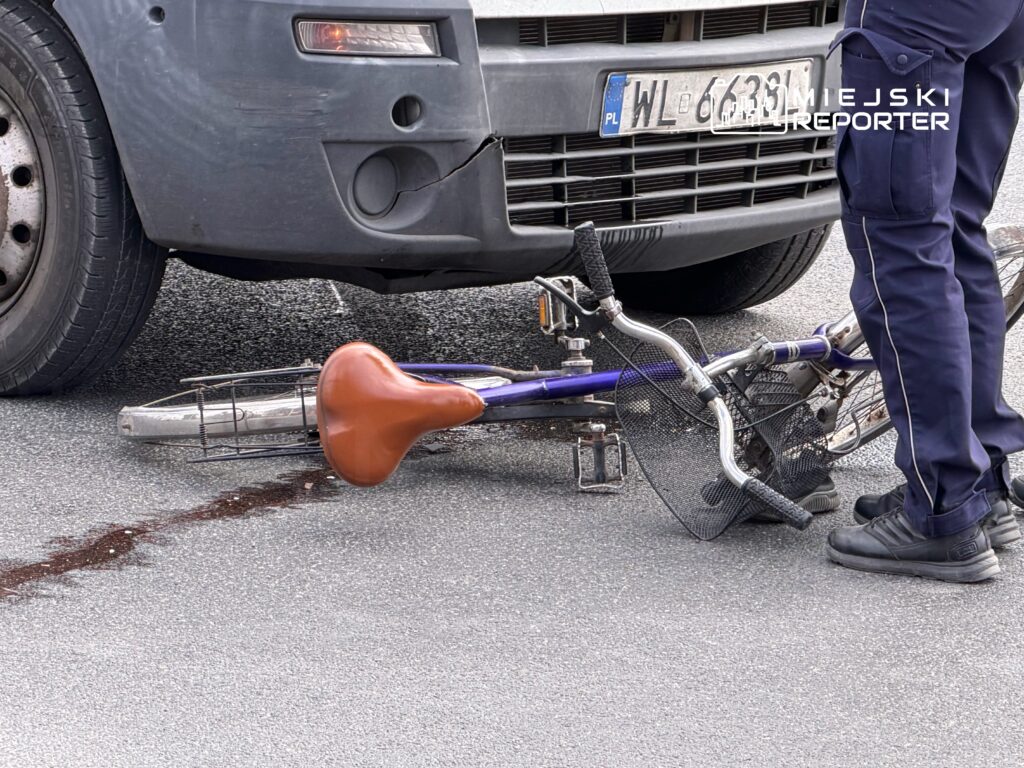 Biały samochód dostawczy stoi obok przewróconego roweru na drodze, a obok stoi policjant w mundurze.