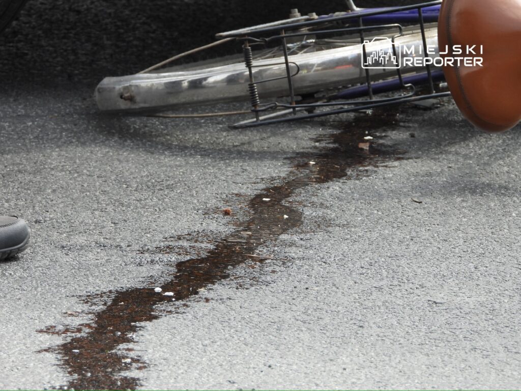 Na asfalcie leży przewrócony rower, obok widoczna plama cieczy, prawdopodobnie oleju.