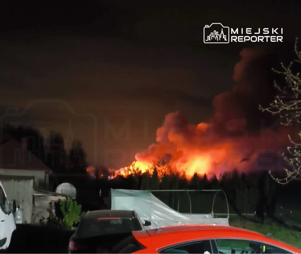 Płonące drzewo z intensywnym ogniem i dymem w tle, widoczne z podwórka z zaparkowanymi samochodami.
