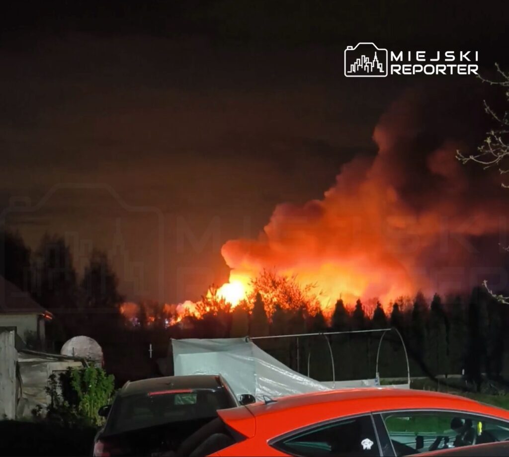 Intensywne płomienie i dym wydobywające się z lasu w nocy, w tle widoczne samochody zaparkowane na podjeździe.