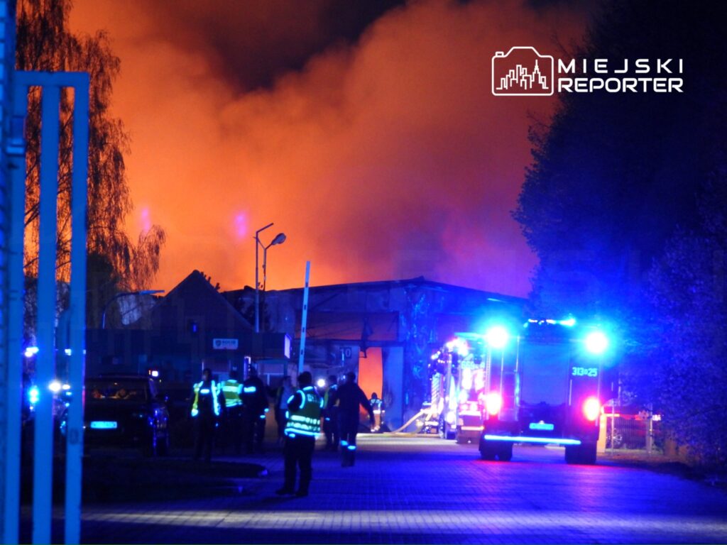 Strażacy w mundurach z odblaskowymi elementami gaszą pożar w budynku, w tle widoczne płomienie i dym.